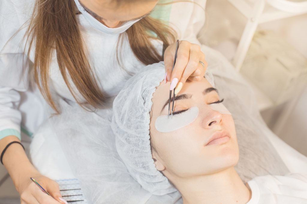 the procedure eyelash extensions for lady girl.cosmetologist glues the eyelashes to the eyelashes a client, the salon is white, girl in med hat